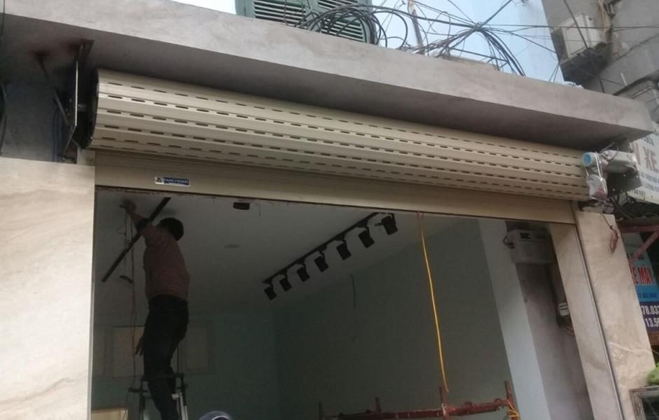 Thợ làm việc dưới cuộn cửa cuốn nhôm khe thoáng màu kem vừa lắp ở mặt tiền cửa hàng