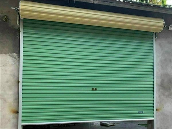 Cửa cuốn tấm liền màu xanh lá đóng gần sát đất, lô cuốn màu kem nằm phía trên ô cửa