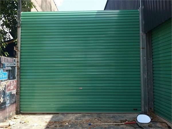 Cửa cuốn tấm liền màu xanh lá khổ rộng đóng kín, bên phải là một bộ cửa cùng màu
