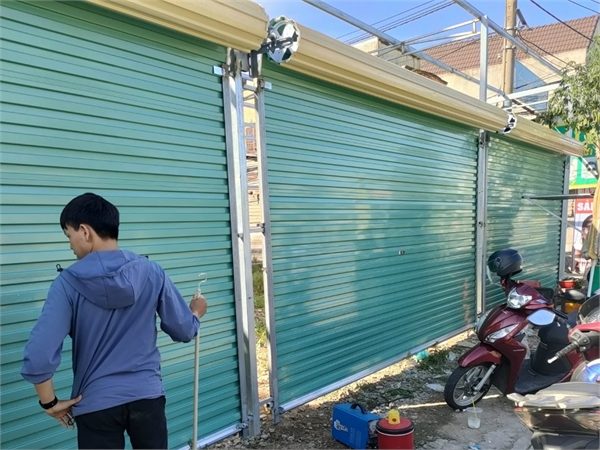 Dãy cửa cuốn Úc tấm liền màu xanh ngọc, lô cuốn màu kem lộ phía trên, một người đứng cạnh cửa