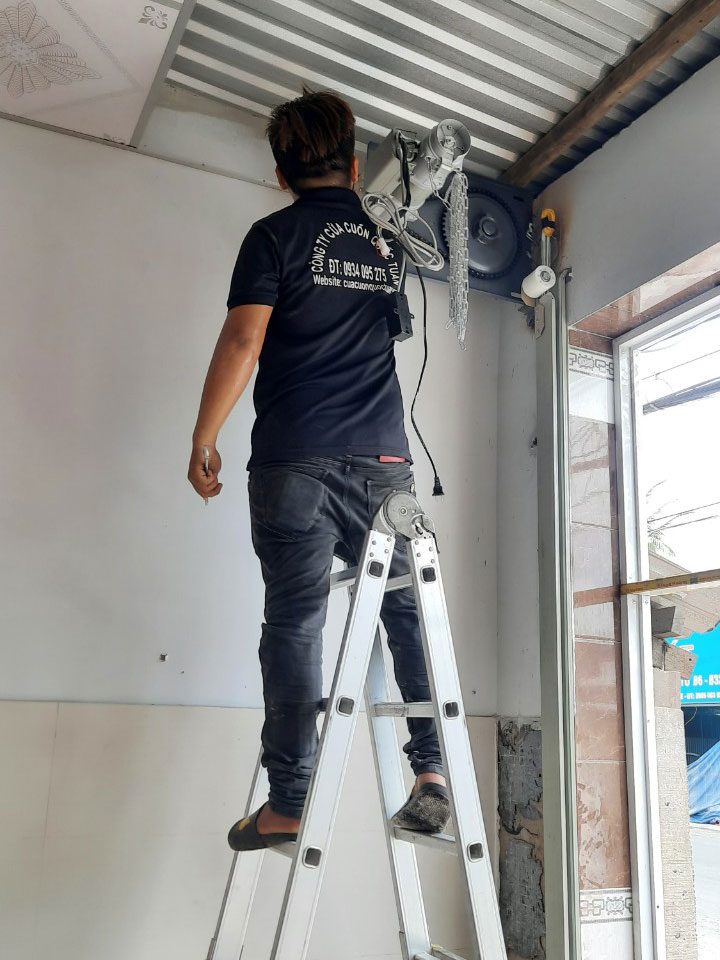 Thợ Quốc Tuấn đứng thang sửa cửa cuốn, kiểm tra motor trên trục cuốn