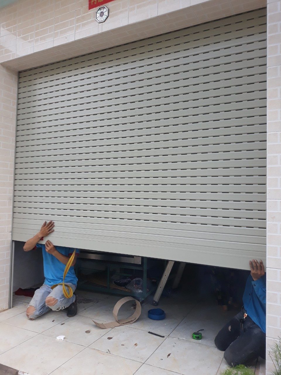 Hai thợ đỡ và căn chỉnh đáy cửa cuốn nhôm khe thoáng màu ghi sáng