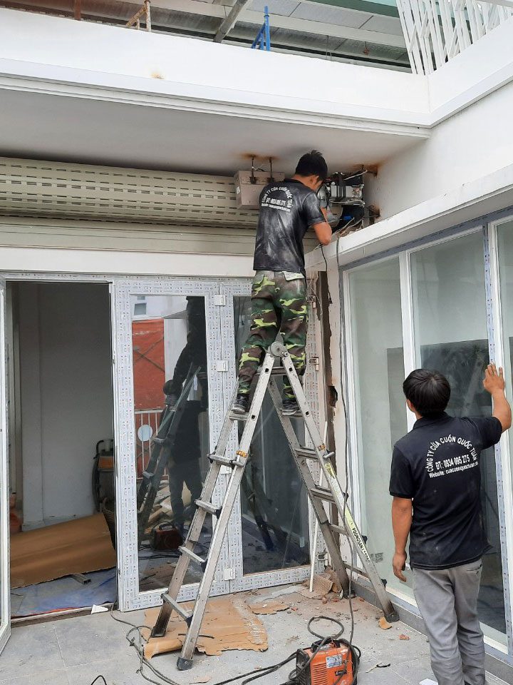 Thợ Quốc Tuấn đứng thang sửa motor cửa cuốn phía trên khung cửa kính