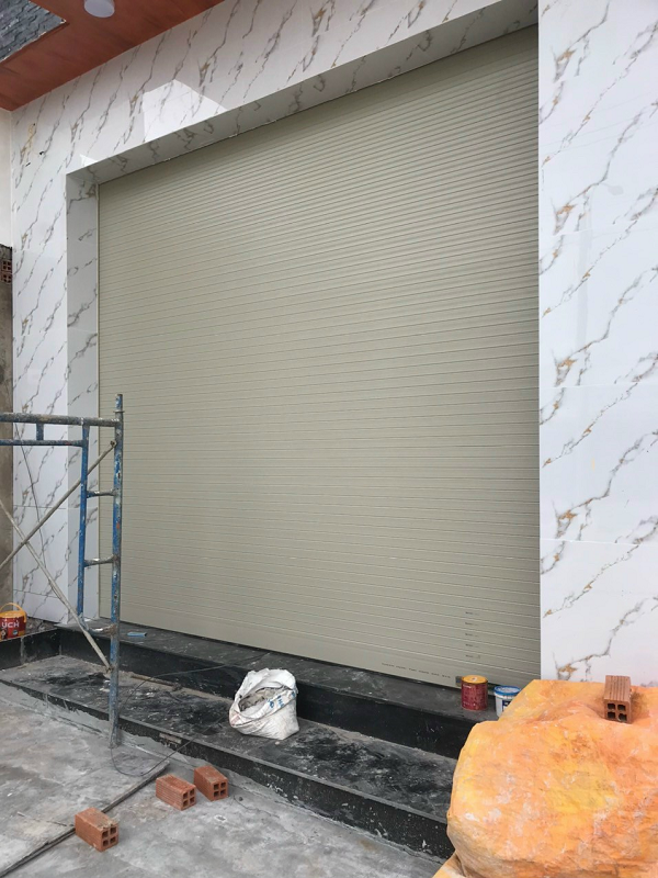 Cửa cuốn lá màu ghi nhạt đóng kín ở mặt tiền nhà ốp đá vân trắng, giàn giáo còn dựng bên trái