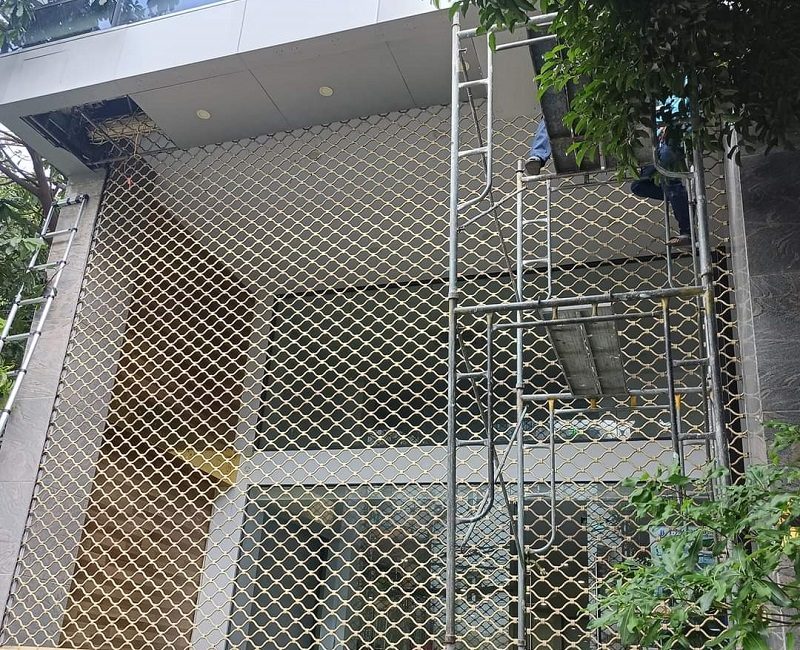 Thợ đứng trên giàn giáo lắp cửa cuốn mắc võng màu vàng kem cho ô cửa cao ở mặt tiền một tòa nhà
