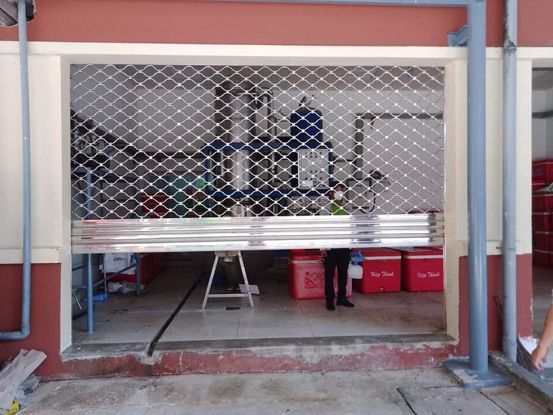 Cửa cuốn mắc võng inox hạ một phần, nhìn xuyên qua lưới thấy máy móc và thùng hàng bên trong