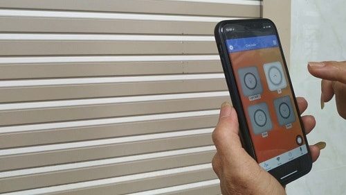 Tay cầm điện thoại mở ứng dụng điều khiển cửa cuốn thông minh, phía sau là cửa cuốn lá màu kem