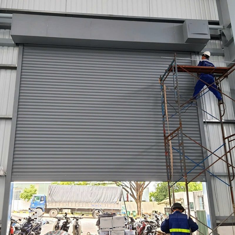 Cửa cuốn khổ lớn màu ghi đang lắp trong nhà xưởng, một thợ đứng trên giàn giáo, một thợ đứng dưới