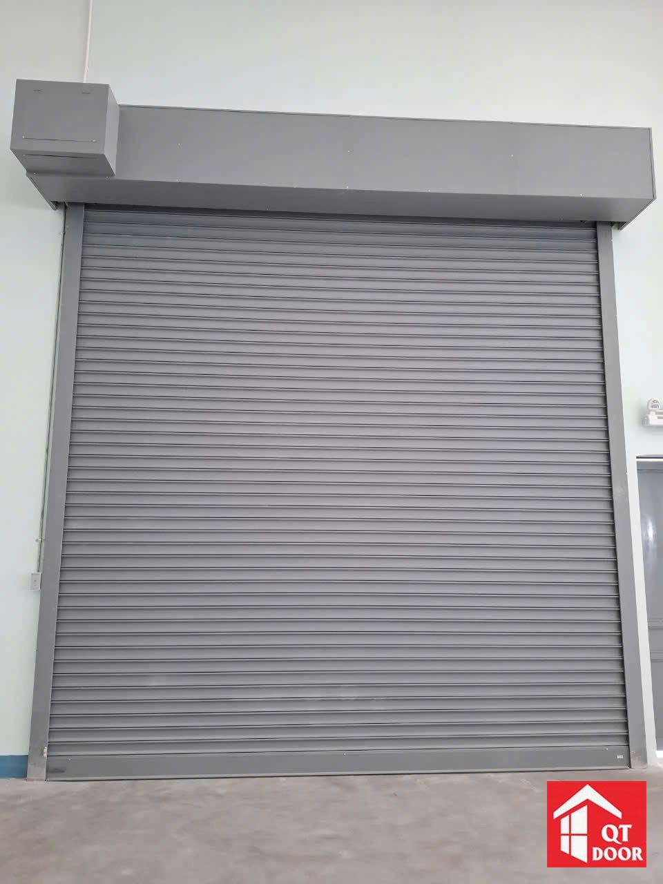 Cửa cuốn màu ghi xám có hộp che lô cuốn phía trên, lắp trong nhà xưởng tường trắng