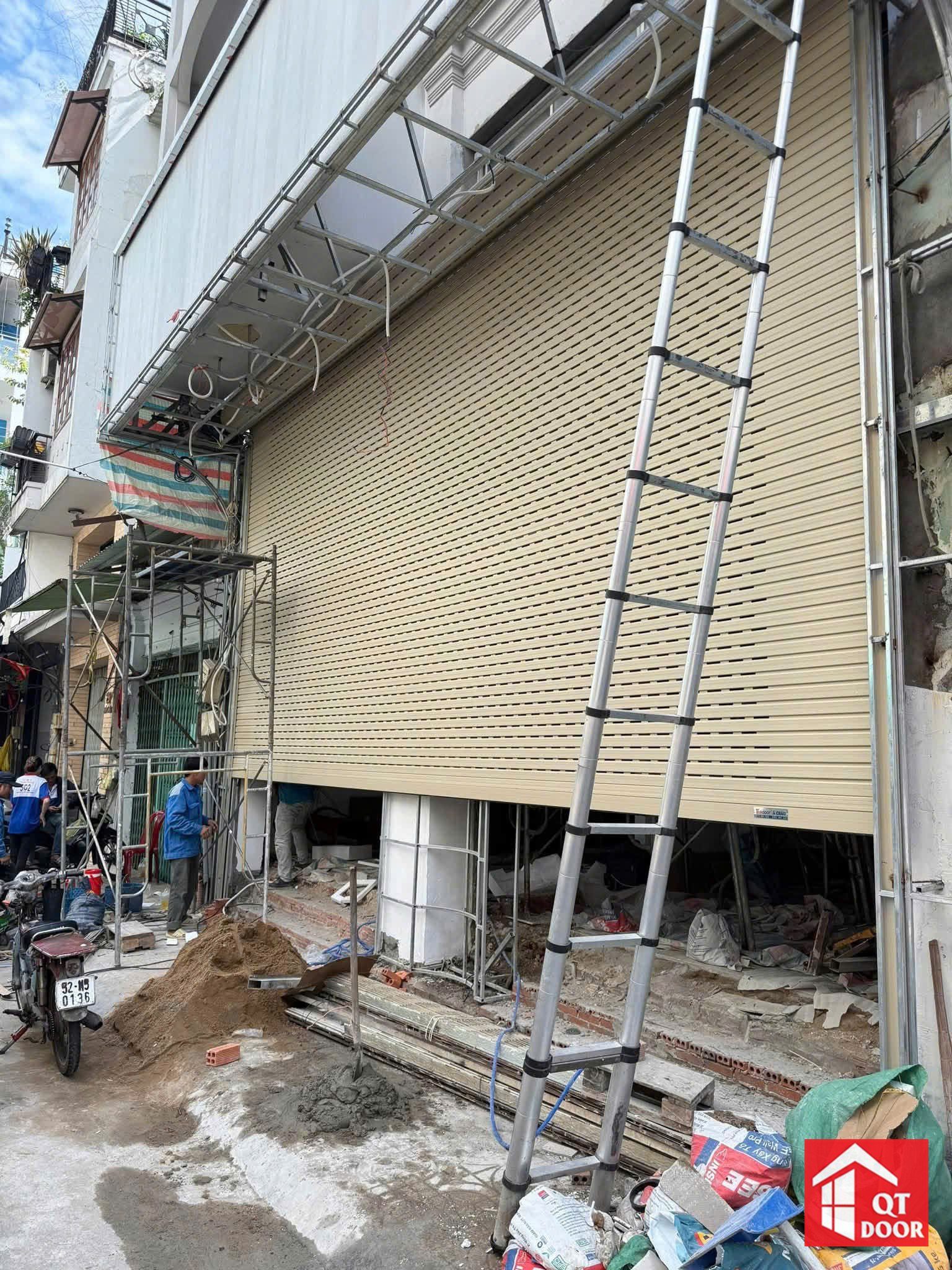 Cửa cuốn khe thoáng màu vàng kem khổ lớn ở mặt tiền nhà phố, thang nhôm và giàn giáo còn dựng