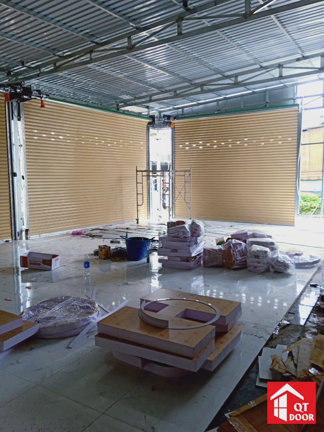 Hai cửa cuốn khe thoáng màu vàng kem lắp ở góc nhà mái tôn, sàn còn vật tư