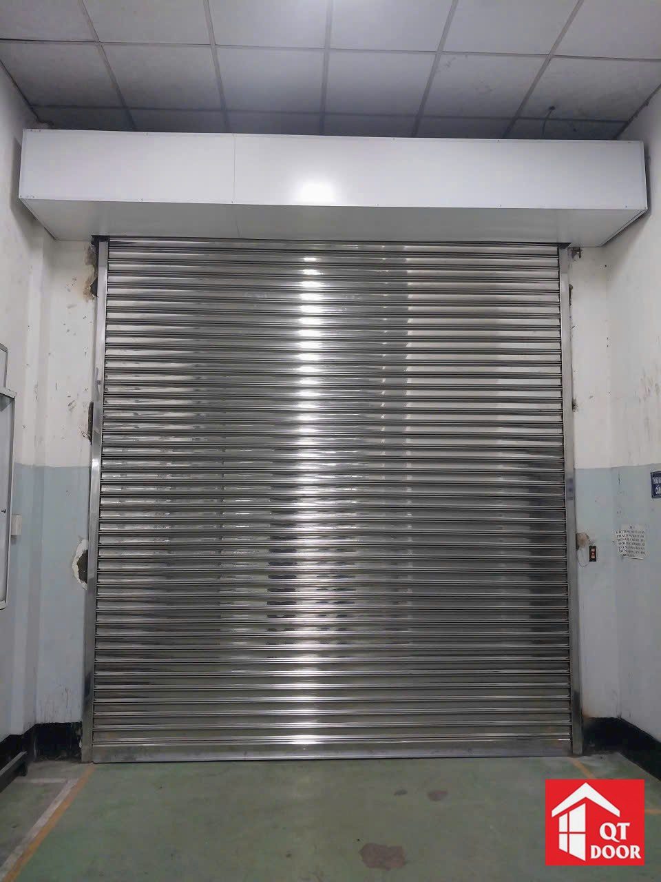 Cửa cuốn inox sáng bóng có hộp che lô cuốn, lắp ở lối vào tường trắng
