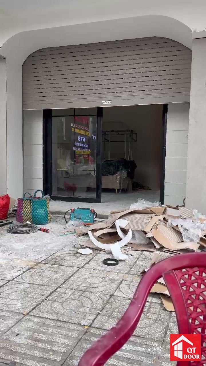 Cửa cuốn màu ghi đang hạ xuống trước mặt tiền cửa kính của một nhà phố mới hoàn thiện