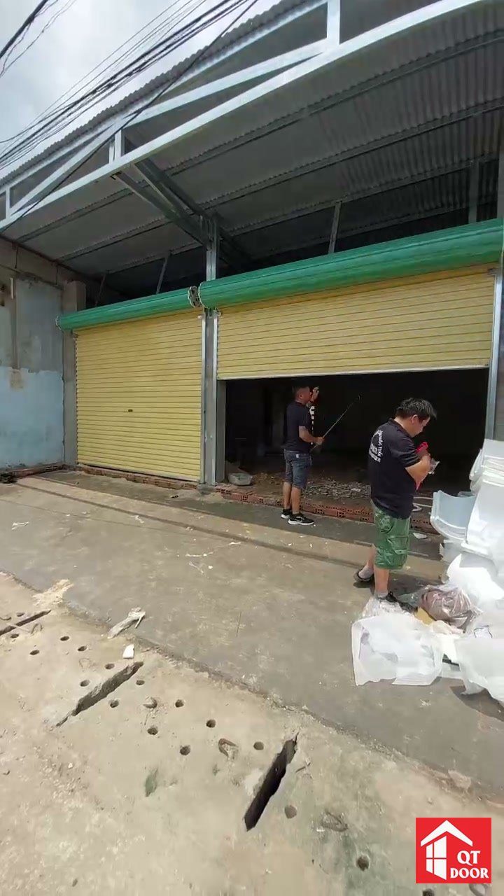 Thợ lắp dãy cửa cuốn màu vàng ở dãy nhà mái tôn, một bộ đang mở